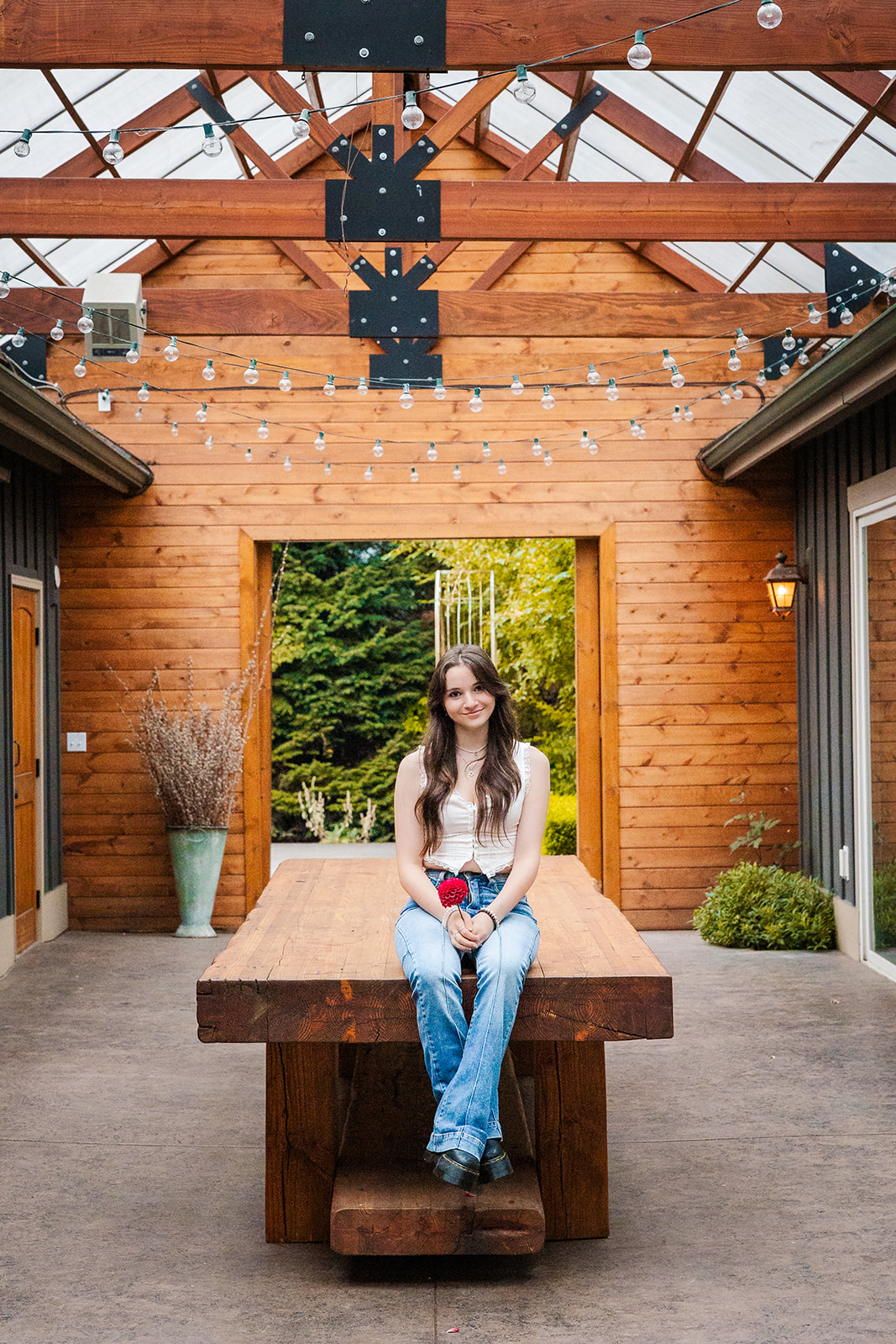 A happy high school senior in jeans and a white blouse sits on a farmtable in a greenhouse holding a red flower