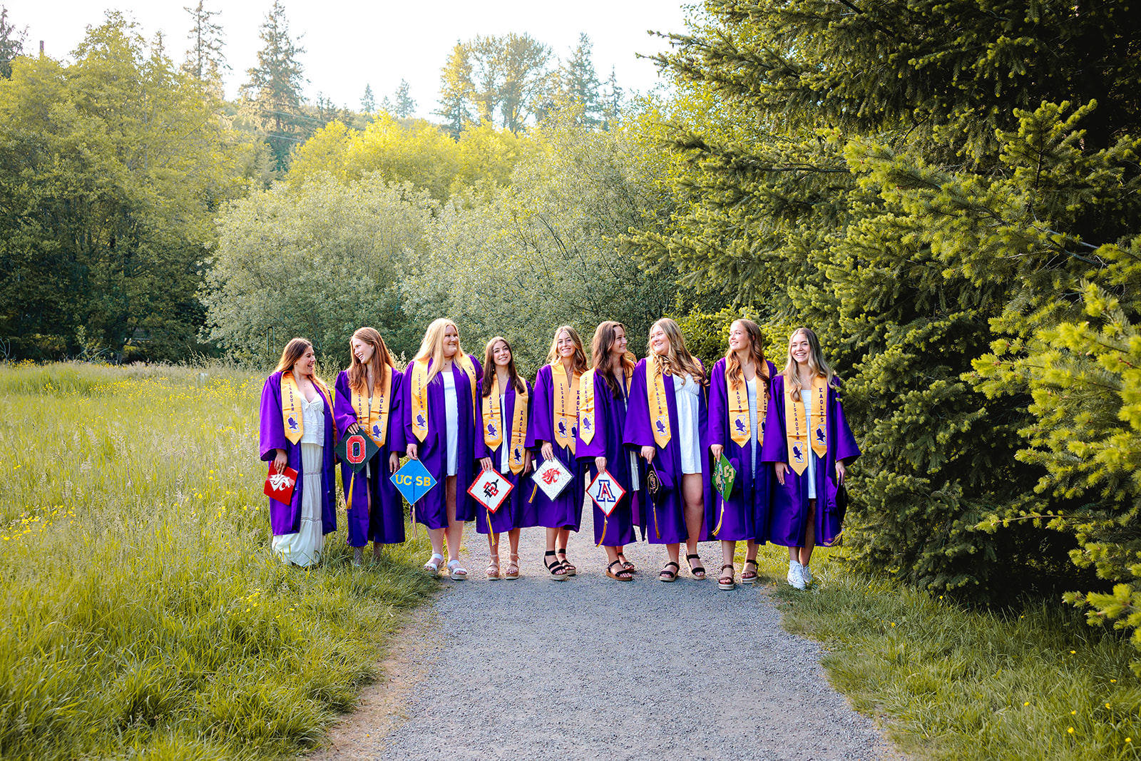A group of high school graduate girls walking in a park path in their robes with caps decorated for their colleges