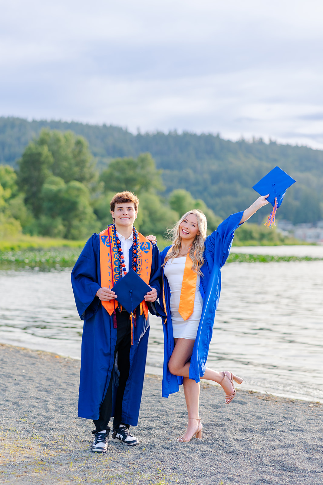 Happy high school seniors in their blue robes smile on a lakefront beach before attending Grad Nights Seattle