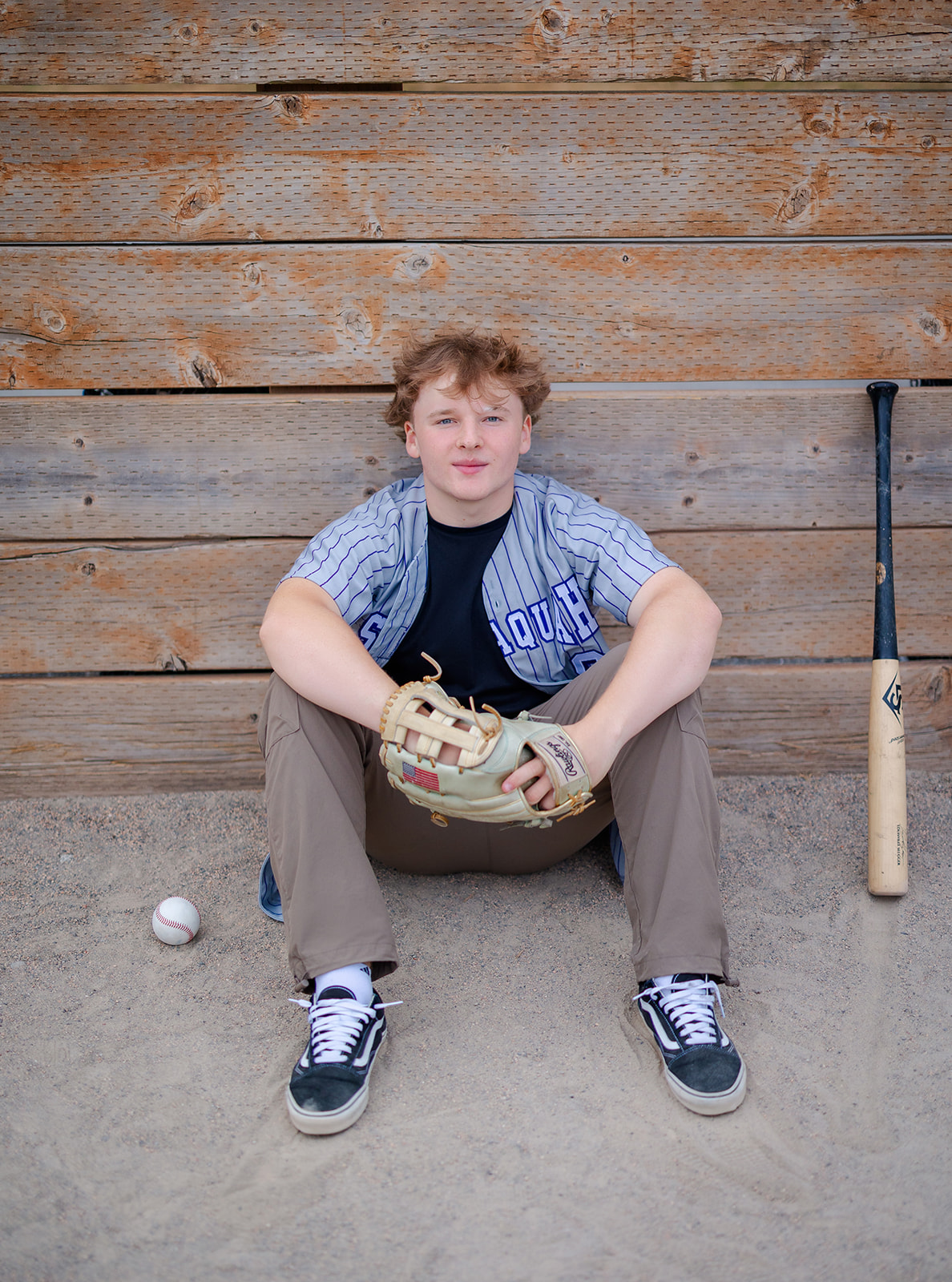 A high school baseball player sits in his jersey and khaki pants with a ball glove and bat against a wooden wall after finding high school internships in Seattle