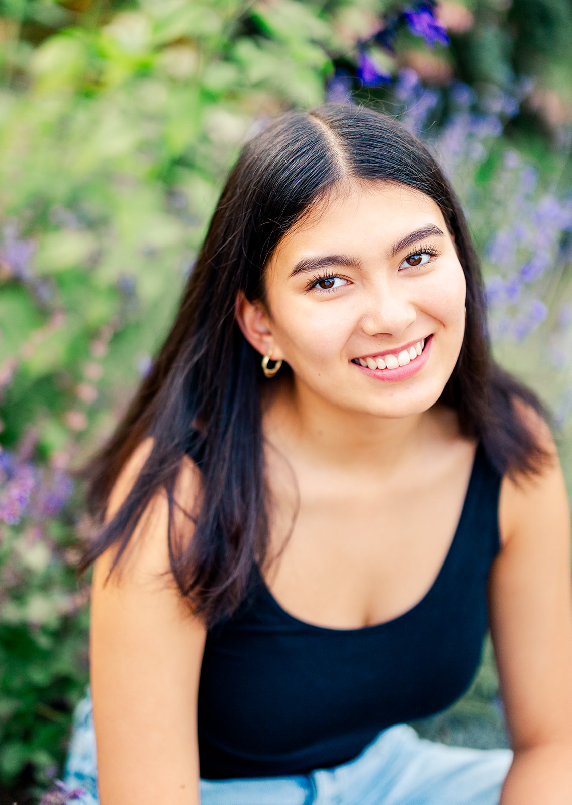 A teenage girl in a black shirt sits in a bed of flowers smiling