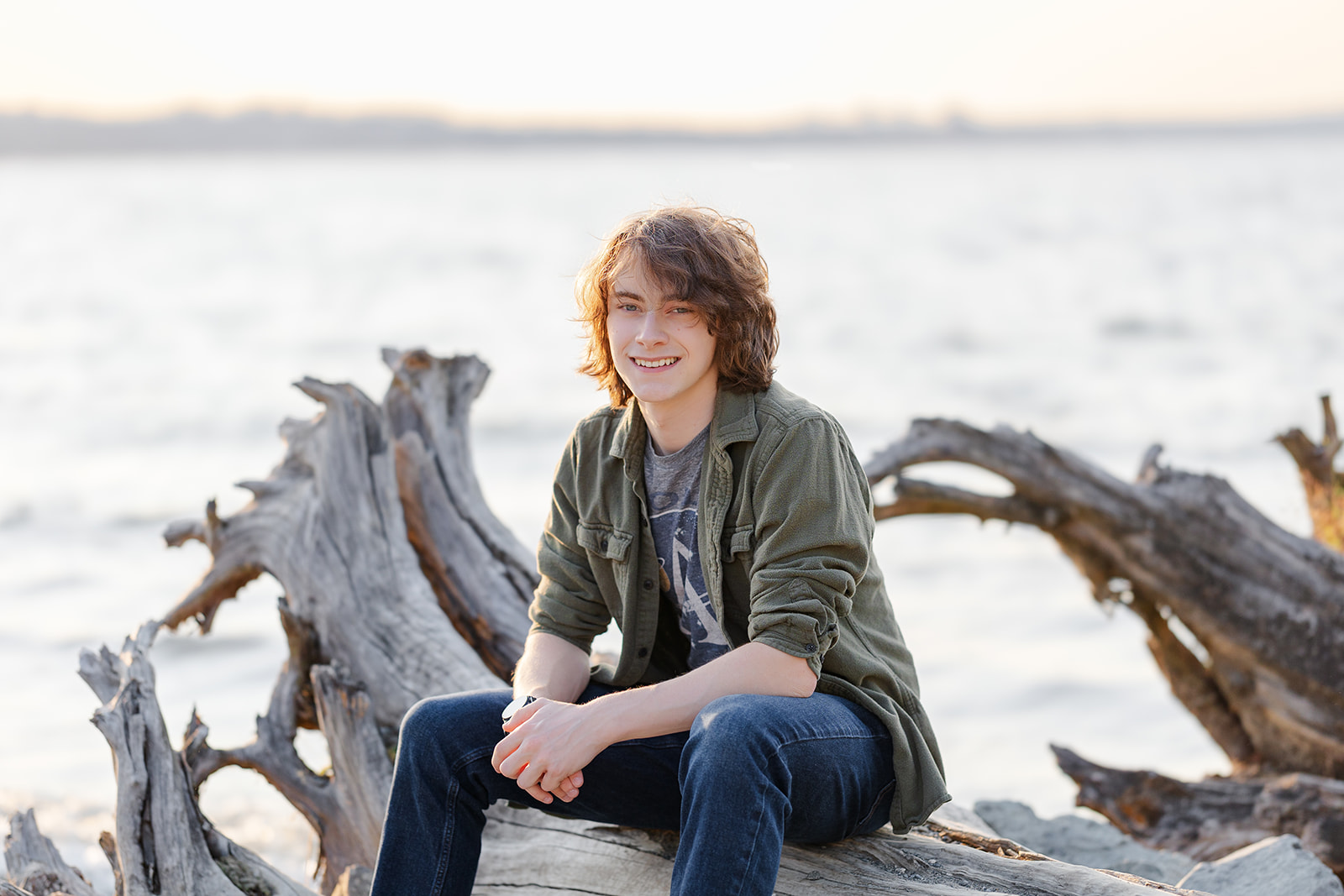 A teenager in an open green shirt and jeans sits on driftwood on the water smiling at sunset after using running start program Seattle