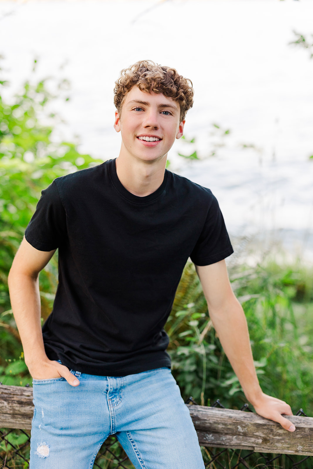 A high school senior in a black shirt leans on a wooden fence smiling after using running start program Seattle