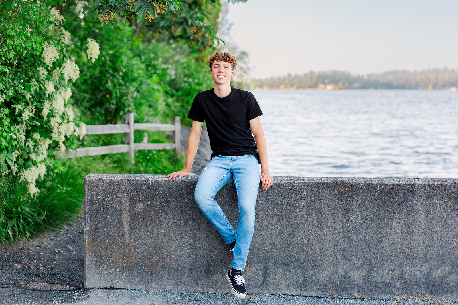 A teenager in jeans and black shirt sits on a waterfront park baricade
