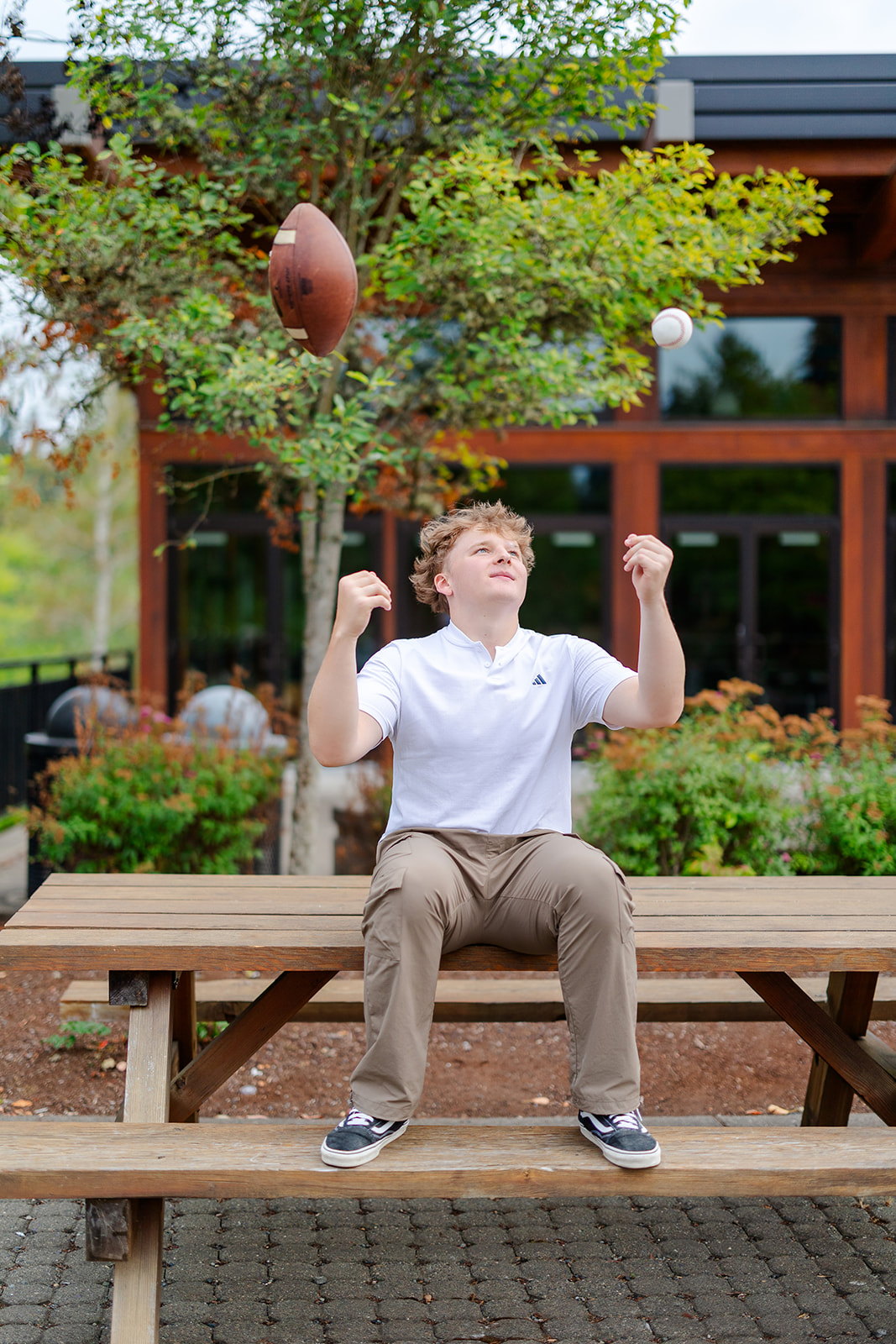 A high school sports player sits on a picnic table tossing a football and baseball in the air after winning scholarships in Seattle