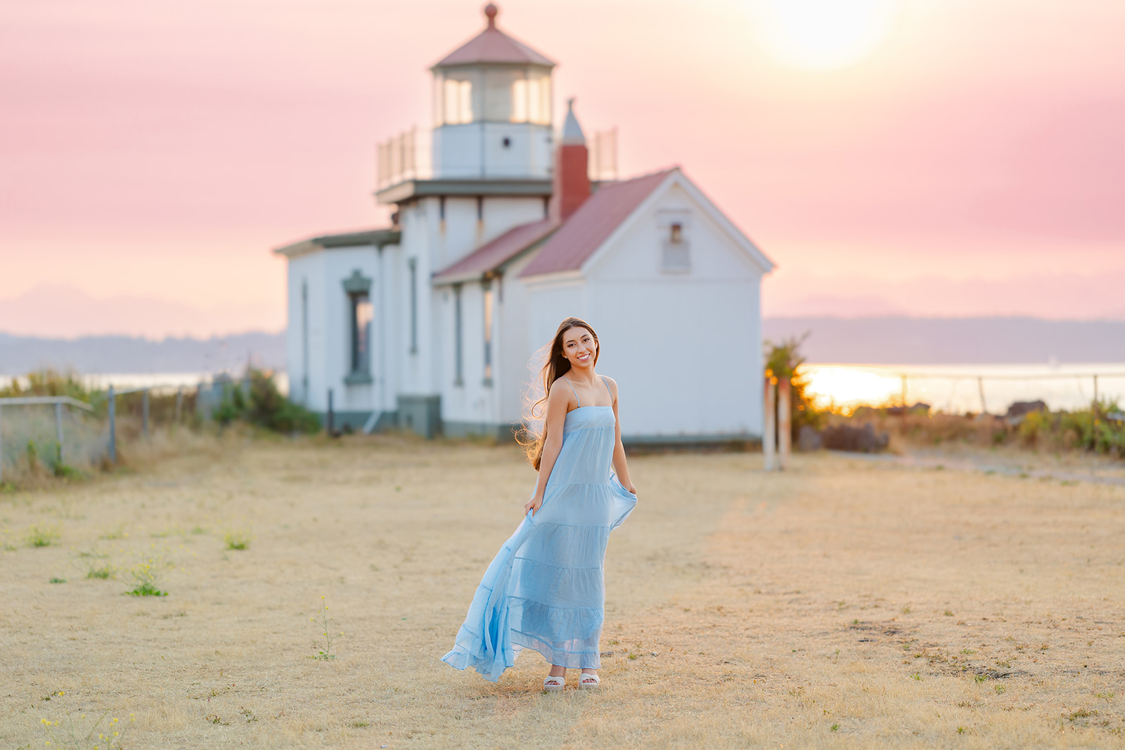 A happy high school senior in a blue dress walks in a lighthouse park at sunset
