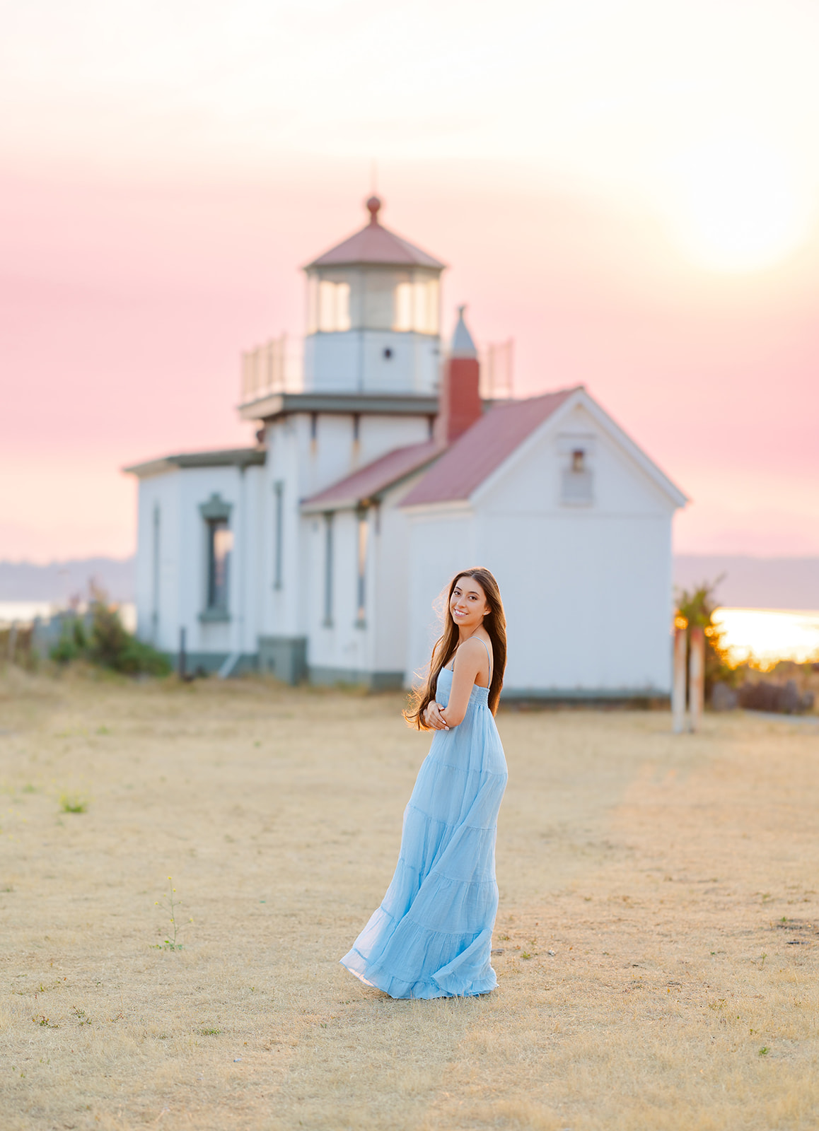 A high school senior walks by a lighthouse at sunset in a flowing blue gown while smiling after being a volunteer on Mercer Island