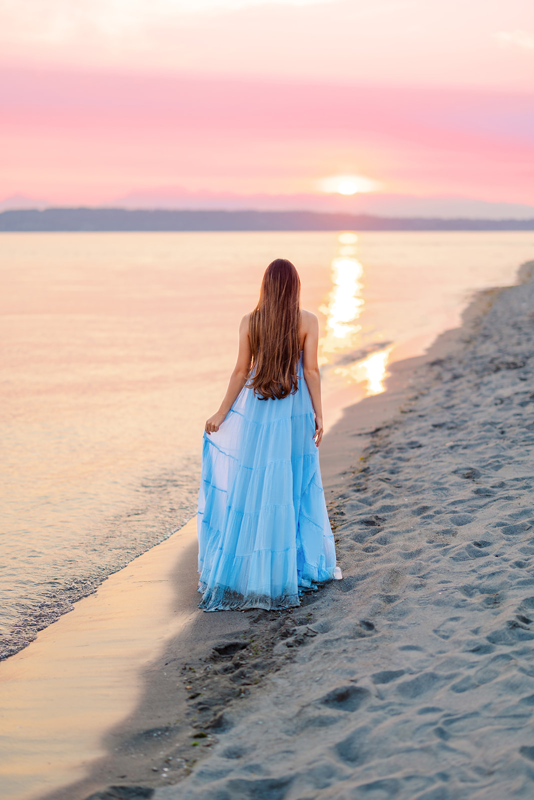 A high school senior in a blue dress walks on the beach holding her dress at sunset after being a volunteer on Mercer Island