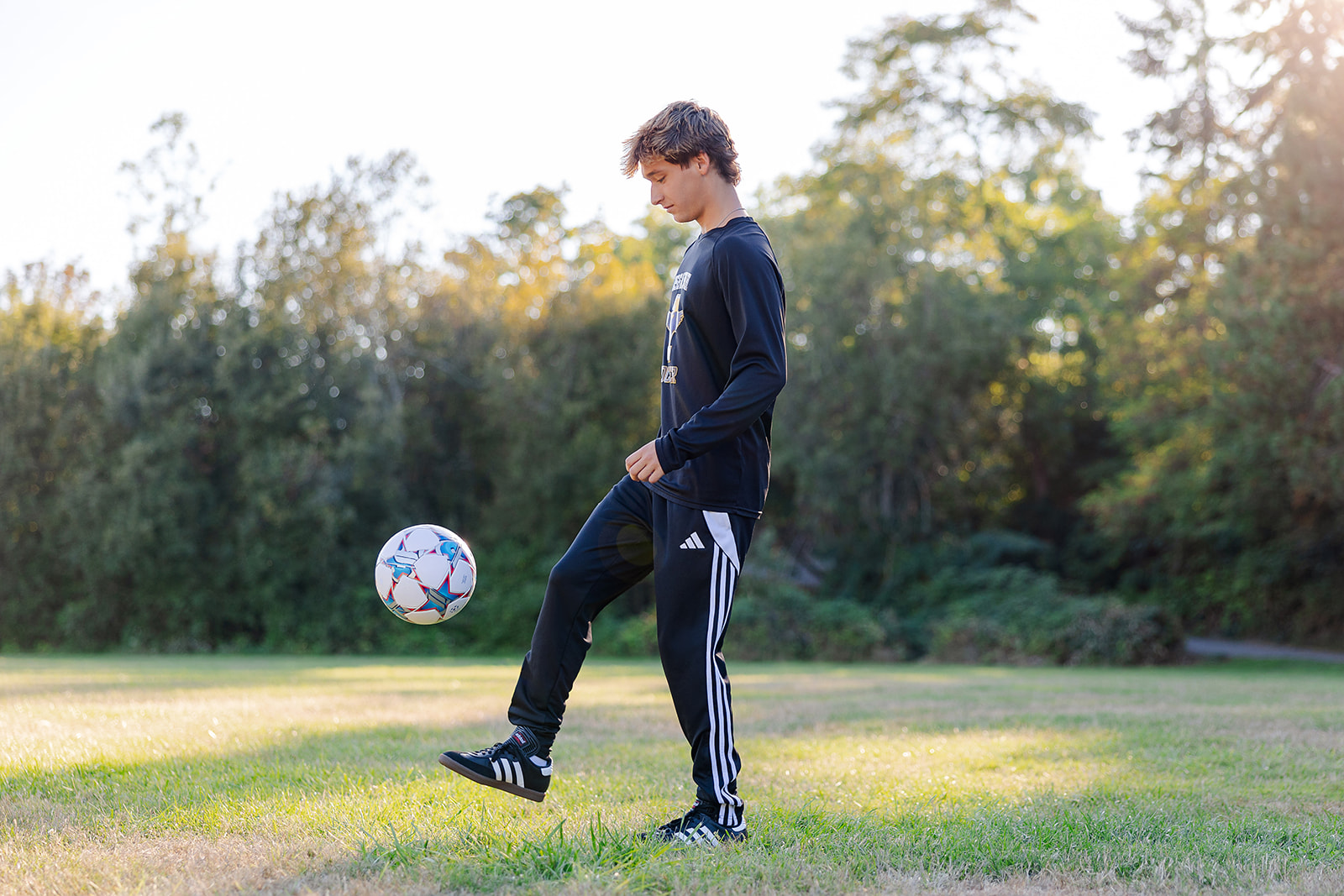 A soccer player juggles a ball in a park field at sunset in black sweatpants and hoodie