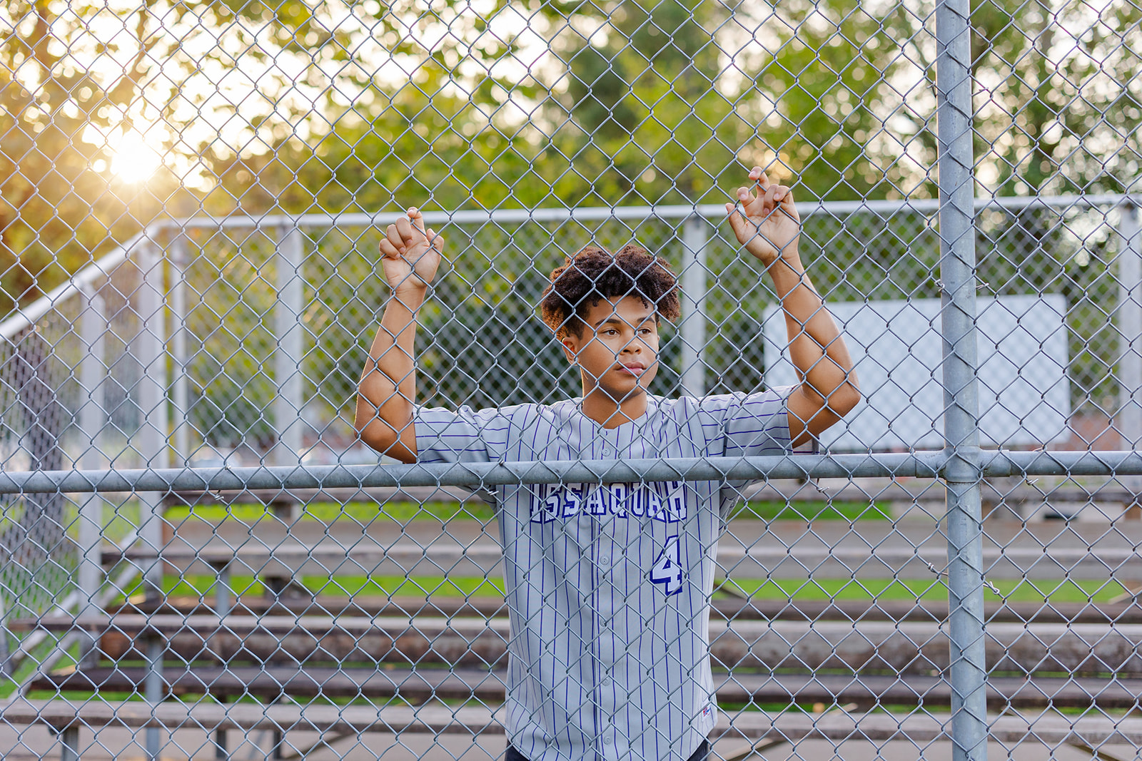 A high school baseball player leans on the backstop fence by the stands in a baseball field at sunset