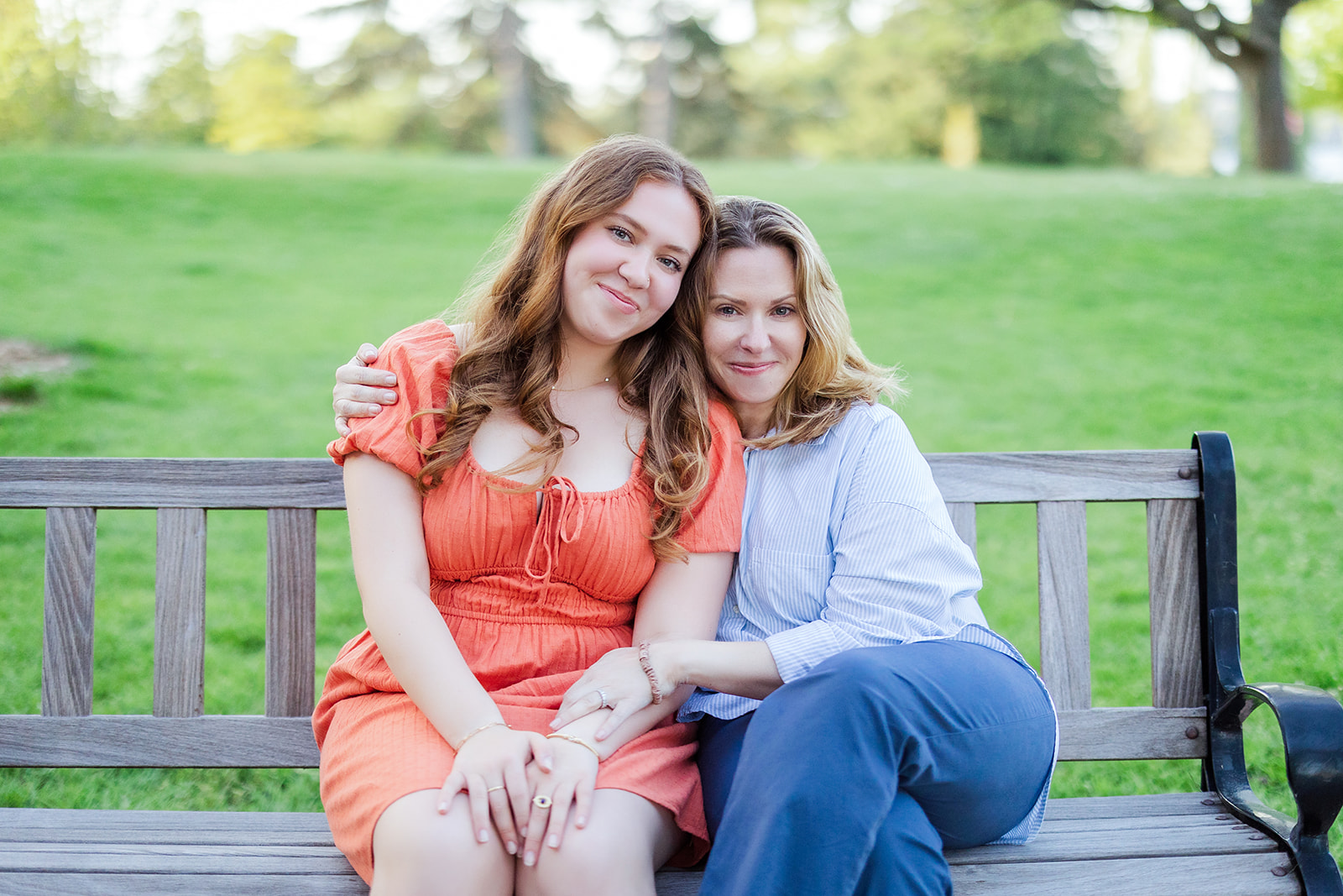 A proud mom sits on a park bench hugging her high school senior daughter in a red dress