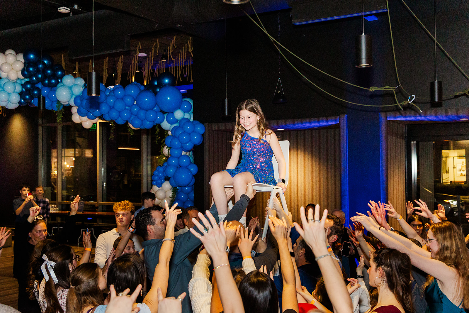 A young girl in a blue dress in lifted in a chair by her guests at her mitzvah
