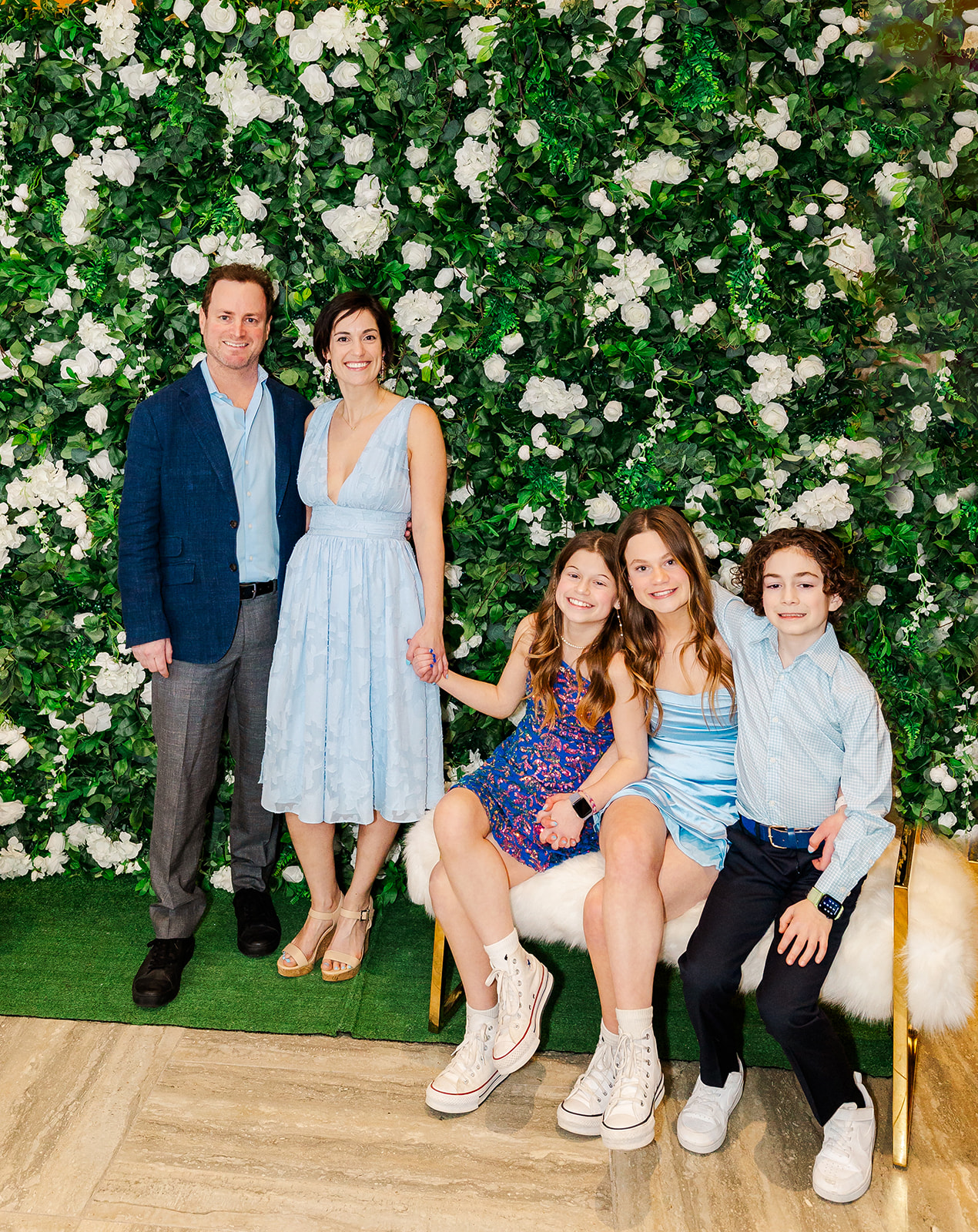 Happy mom and dad stand in front of a flower wall behind their three young children smiling on a bench